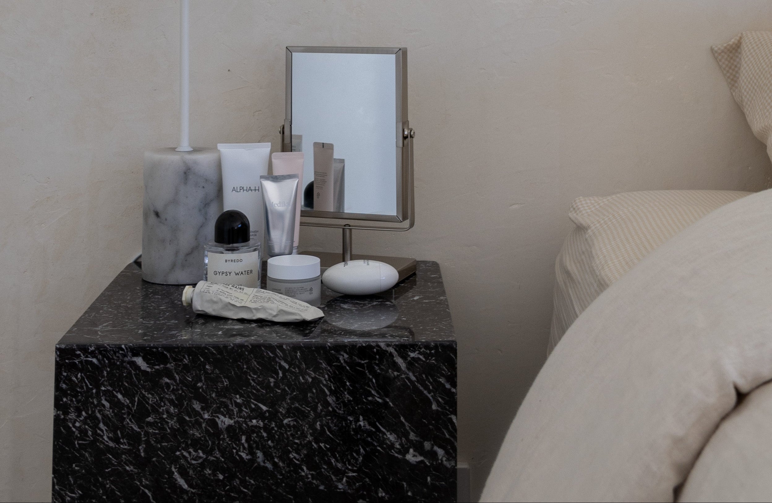 Nightstand with skincare products and a mirror next to a bed in a bedroom.